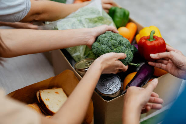 Several hands placing food items into a brown cardboard box for donation.